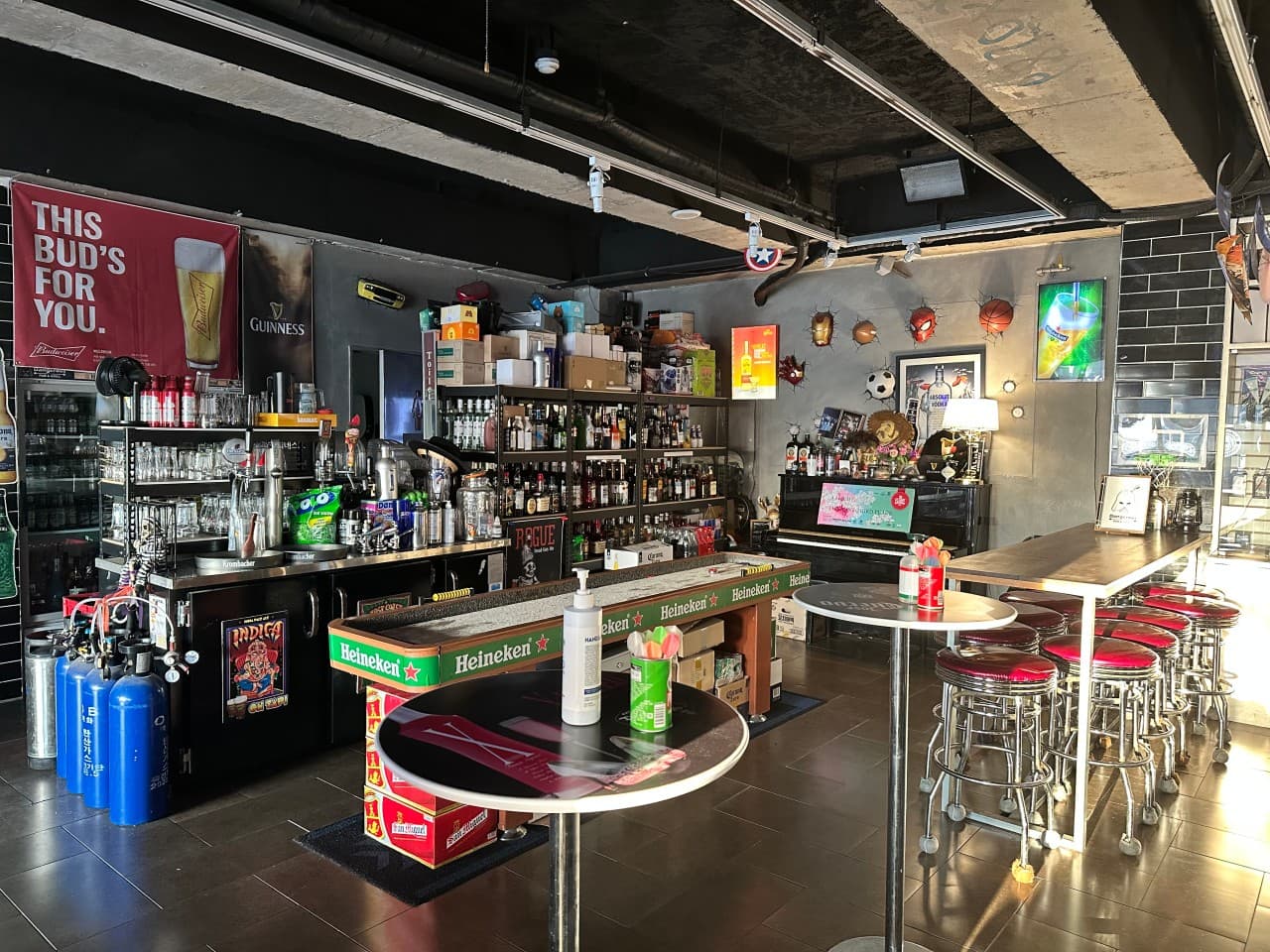 충청북도 충주시 DANGEROUS PUB 바(Bar) - 붉은 스툴과 높은 테이블이 배치된 실내. 벽면 주류와 캐릭터 장식, 자연광이 비치는 창가 쪽 1인 좌석에서 혼술 상황을 연출함.