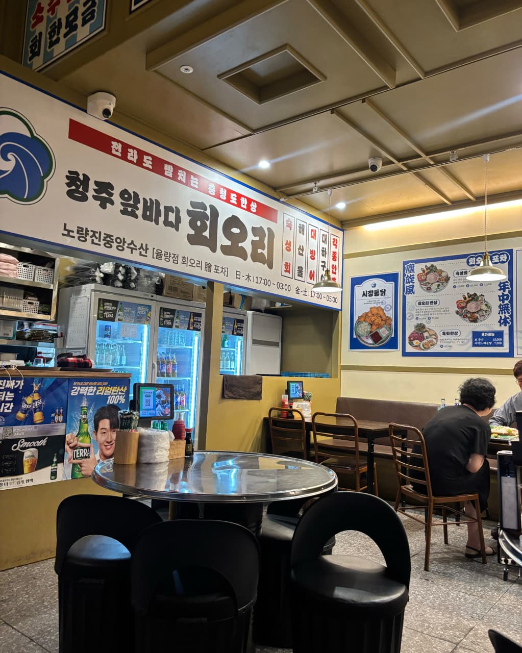 충청북도 청주시 요리주점 회오리 청주율량점의 노란 조명 아래 스테인리스 원형 테이블과 1인 다찌석. 태블릿 주문기가 설치된 좌석에서 혼술을 즐기는 상황.