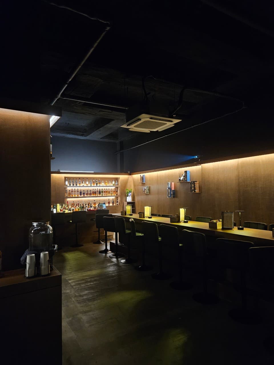 충청남도 천안시 혼술바 고도 불당점 바(Bar) 내부. 어두운 천장 아래 긴 다찌석과 1인용 스탠드 조명이 배치되어 있고 벽면 선반에 다양한 위스키 병이 진열됨.