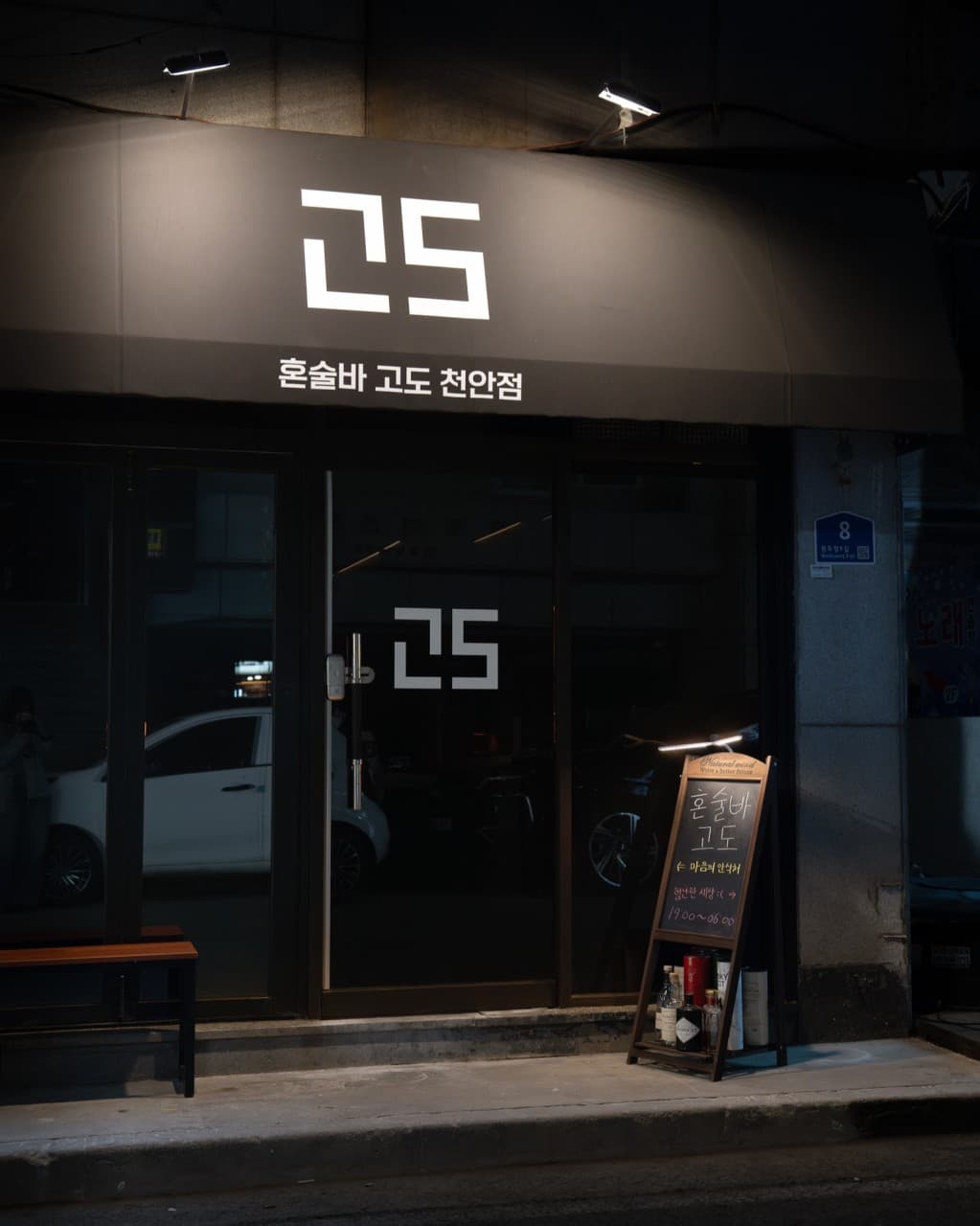 충청남도 천안시 혼술바 고도 천안점 외부 전경. 어두운 밤 조명이 비치는 검은색 어닝과 메뉴가 적힌 입간판이 놓인 1인 바 입구의 정돈된 모습.