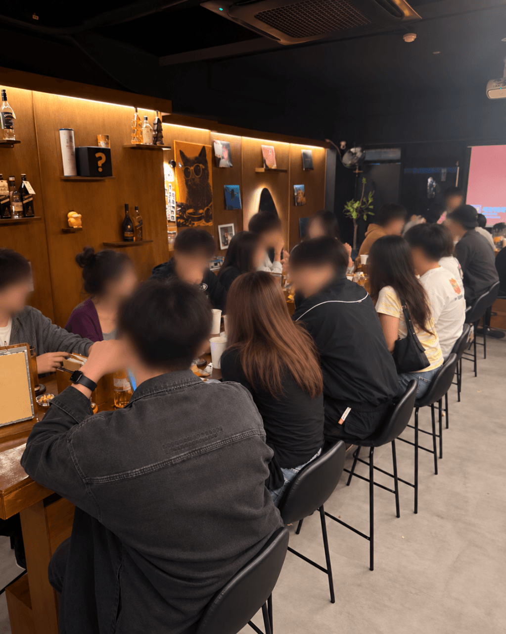 충청남도 천안시 혼술바 고도 천안점 바(Bar)의 노란 간접 조명이 비치는 나무 선반 아래 긴 다찌석. 1인석에서 잔에 담긴 술과 함께 혼술을 즐기는 사람들의 뒷모습.
