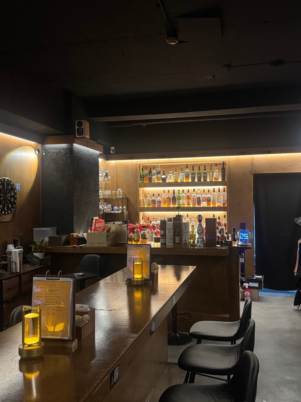충청남도 천안시 혼술바 고도 천안점 바(Bar) 내부의 긴 나무 다찌석과 1인용 좌석들. 선반 위 다양한 술병을 비추는 간접 조명과 테이블마다 놓인 램프가 연출하는 1인 혼술 전용 공간.