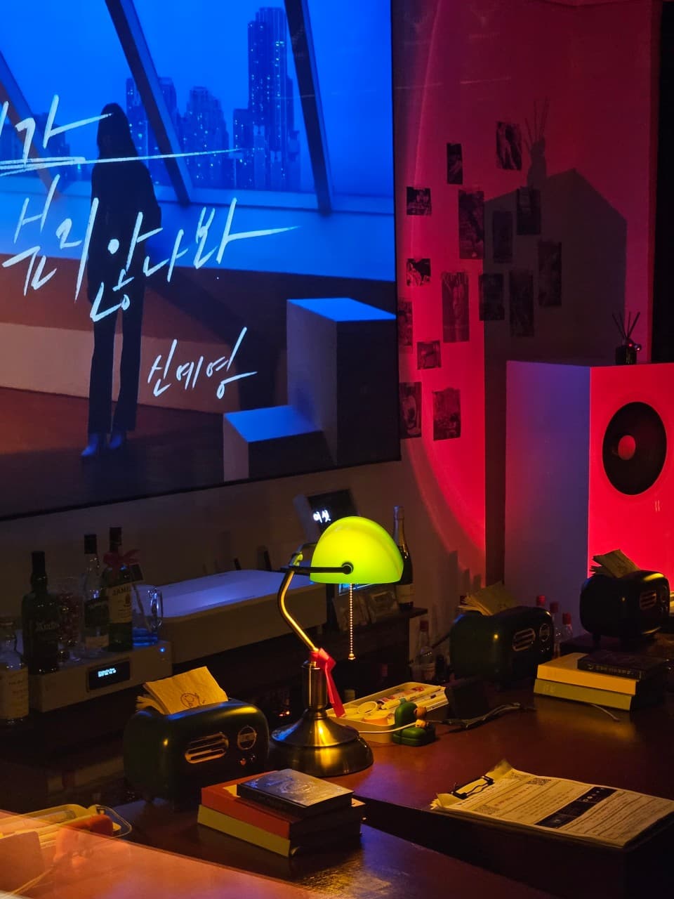 충청남도 천안시 불당동골방 1인 혼술 공간. 초록색 스탠드 조명이 켜진 다찌석과 대형 스크린. 붉은 조명 아래 책과 기타 소품이 배치된 모습.