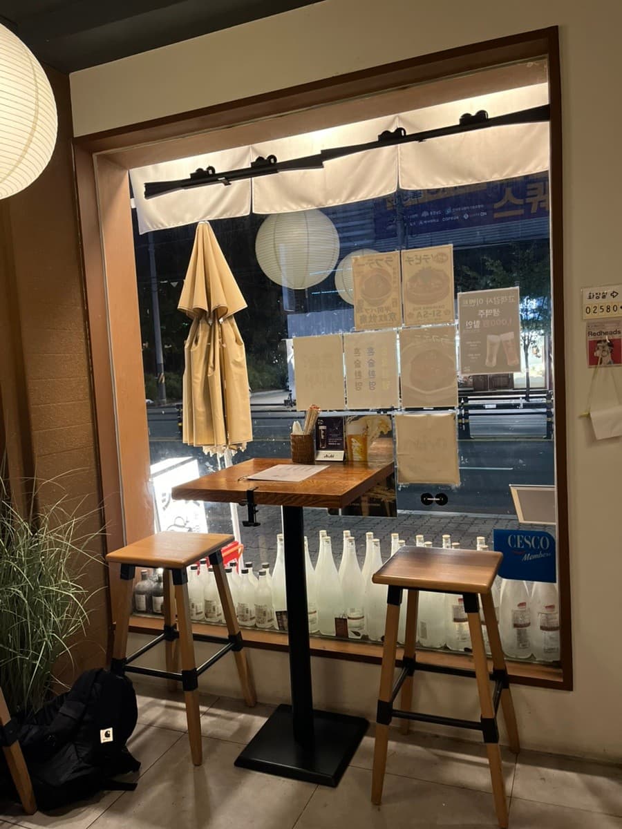 충청남도 천안시 오키나와펍시사 이자카야 창가 다찌석. 둥근 조명 아래 나무 테이블과 의자가 배치된 1인 혼술 공간. 유리창에 붙은 메뉴판과 바닥에 진열된 술병들이 보임.