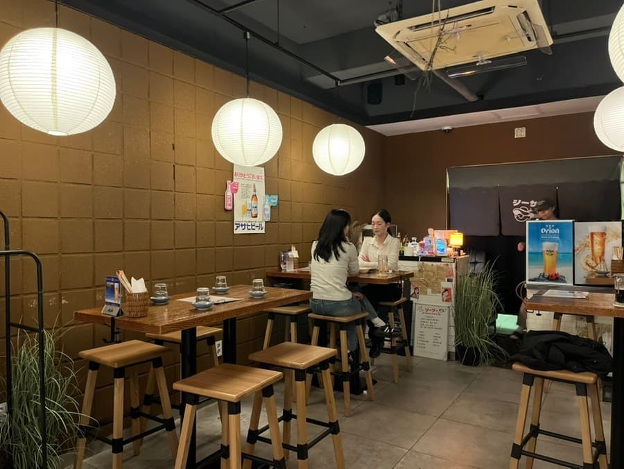 충청남도 천안시 이자카야 오키나와펍시사 내부. 둥근 펜던트 조명 아래 높은 목재 테이블과 1인 혼술용 다찌석 의자가 배치된 실내 공간.