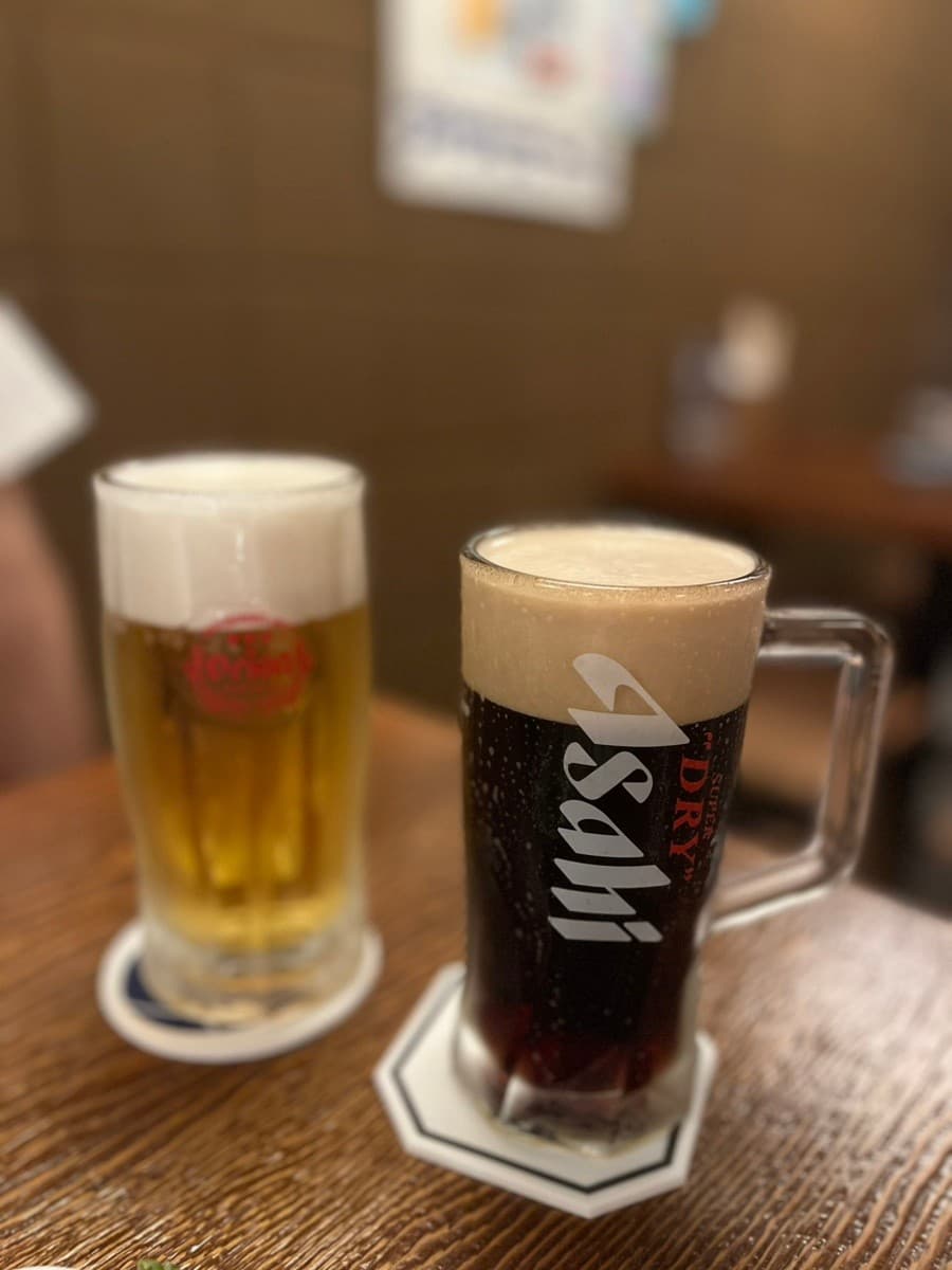 충청남도 천안시 이자카야 오키나와펍시사 나무 테이블 위 아사히 흑맥주와 오리온 생맥주. 조명 아래 혼술 다찌석에서 즐기는 거품 가득한 맥주.